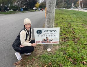 Alexis from Bottaro Law Firm squatting next to a Bottaro Law Firm lawn sign with the street in the background