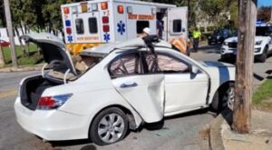 exterior of white car on road on sunny day with trunk open and smashed passenger door with emergency vehicle in the background