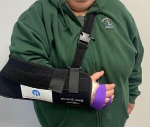 close-up of a person wearing a green hooded sweatshirt with right arm in a black sling and purple medical tape over four fingers